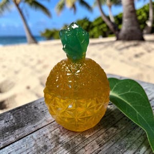 May include: A translucent yellow pineapple-shaped object with a textured, diamond-patterned surface and a solid green top. The object is crafted from a glossy material that catches the light, featuring a rounded body and a pointed, leafy crown.