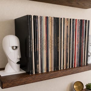 May include: A collection of vinyl records arranged vertically between two white, abstract, geometric head-shaped bookends. The records feature various spine colors and text, including the visible title Pink Floyd The Dark Side of the Moon.