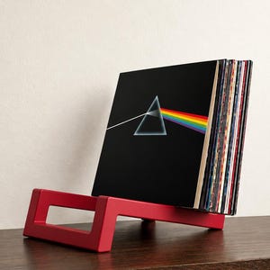 Può includere: Un supporto rosso per dischi in vinile che contiene una collezione di dischi, tra cui uno con una copertina nera raffigurante un triangolo bianco e un prisma a spettro arcobaleno, l'iconica grafica dell'album The Dark Side of the Moon.