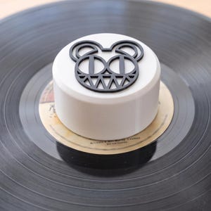 Op de afbeelding: Een wit cilindrisch platengewicht met een zwarte afbeelding van een gestileerd berengezicht met scherpe tanden aan de bovenzijde, centraal geplaatst op een zwarte vinylplaat met een beige label met kleine gedrukte tekst.