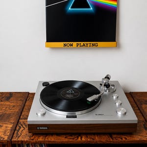 Könnte beinhalten: Ein silberner Yamaha-Plattenspieler mit einer schwarzen Schallplatte steht auf einem Holzschrank. Darüber befindet sich ein schwarzer quadratischer Rahmen mit dem Pink-Floyd-Prisma und einem gelben Regal mit dem Text NOW PLAYING.