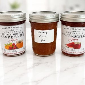 May include: Three glass jars with silver lids containing jam. The left jar is labeled Organic Double Golden Raspberry Jam. The middle jar has a white label with handwritten text reading Strawberry Rhubarb Jam. The right jar is labeled Organic Watermelon Jam.