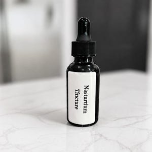 May include: A dark amber glass dropper bottle with a black cap and a white rectangular label. The label features the black printed text Nasturtium Tincture. The bottle has a cylindrical body and a tapered neck with a dropper mechanism on top.