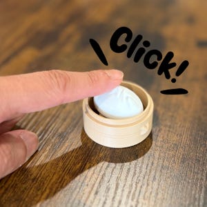 Peut inclure: Un petit panier vapeur de style bambou, de couleur claire, contenant un seul objet blanc de forme ovale ressemblant à une boulette. Le mot Click! est écrit en lettres noires au-dessus de l'objet, sur lequel un doigt appuie.