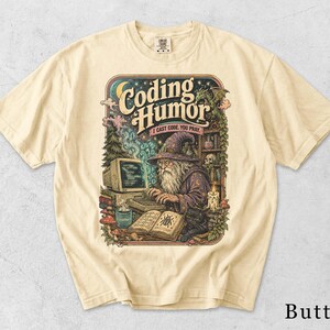 Könnte beinhalten: Ein butterfarbenes T-Shirt mit einer grafischen Illustration eines Zauberers, der an einem Computer tippt. Der Text lautet "Coding Humor" und "I cast code. You pray.". Das Design zeigt einen Drachen, eine schwarze Katze, ein Zauberbuch und eine Kerze.