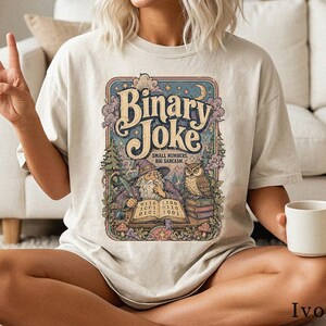 Könnte beinhalten: Ein elfenbeinfarbenes T-Shirt mit einem Grafikdruck eines Zauberers mit Federkiel und einer Eule auf Büchern. Der Text lautet Binary Joke, Small Numbers, Big Sarcasm, mit Binärcode auf einem offenen Buch, umgeben von Bäumen, Wolken und Sternen.