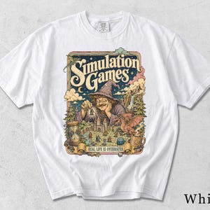 Könnte beinhalten: Ein weißes Kurzarm-T-Shirt mit einem farbenfrohen Grafikdruck eines Zauberers, der ein Brettspiel spielt. Der Text "SIMULATION GAMES" steht oben und "REAL LIFE IS OVERRATED." auf einem Banner unten, umgeben von einem Drachen und einer Burg.