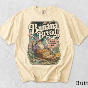 Könnte beinhalten: Ein cremefarbenes Kurzarm-T-Shirt mit einem Grafikdruck eines bärtigen Zauberers mit Hut, der hinter einem Bananenbrot steht. Der Text lautet Banana Bread und Magic You Can Eat. Das Design zeigt zudem eine Burg, Pflanzen und Bananen.