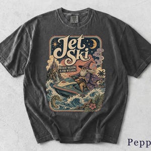 Peut inclure: Un t-shirt gris anthracite avec une impression graphique représentant un magicien sur un jet ski au milieu des vagues. Le texte indique Jet Ski et Because Walking Is For Wizards. L'illustration inclut un château, un serpent de mer et une lune.