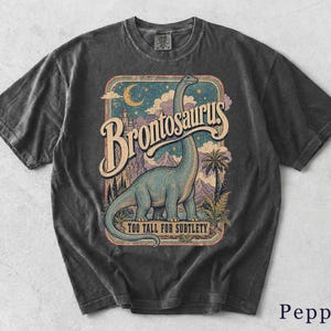 Könnte beinhalten: Ein anthrazitfarbenes T-Shirt mit einem Grafikdruck eines blauen Brontosaurus in einer Landschaft mit Bergen, Palmen und einer Mondsichel. Der Text lautet Brontosaurus und Too Tall For Subtlety in einer Schriftart im Vintage-Stil.