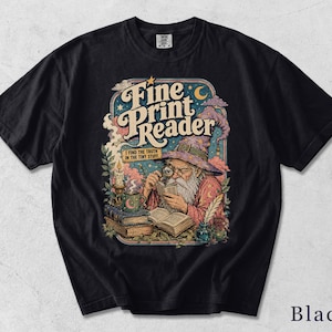 Könnte beinhalten: Ein schwarzes T-Shirt mit einer grafischen Illustration eines Zauberers mit langem weißen Bart und spitzem Hut, der mit einer Lupe in einem Buch liest. Der Text lautet Fine Print Reader und I FIND THE TRUTH IN THE TINY STUFF, mit Kerzen, Büchern und einer Feder.