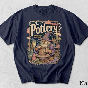 Könnte beinhalten: Ein marineblaues T-Shirt mit einer grafischen Illustration eines Zauberers, der einen Tontopf auf einer Töpferscheibe formt. Das Design zeigt eine Eule, verschiedene Töpferwaren und den Text "Pottery I MAKE MUD LOOK GOOD." Der Zauberer trägt eine violette Robe.