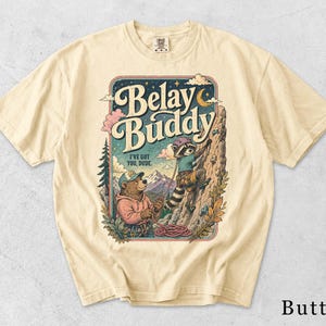 Könnte beinhalten: Ein butterfarbenes Kurzarm-T-Shirt mit einer Grafik von einem Bären und einem Waschbären beim Klettern. Der Text lautet Belay Buddy und I've got you, dude. Die Illustration zeigt Berge, Bäume, einen Halbmond und eine Kletterausrüstung.