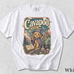 Könnte beinhalten: Ein weißes T-Shirt mit einem Grafikdruck eines Cavapoo-Hundes, der als Zauberer in einem violetten, mit Sternen gemusterten Umhang und Hut gekleidet ist. Das Design enthält den Text "Cavapoo" und "Part dog, part teddy bear, all wizard," einen Zauberstab.
