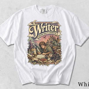 May include: A white short-sleeve t-shirt featuring a colorful graphic print of a person writing at a desk with a quill, surrounded by books, a dragon, a castle, and a cat. The text reads Writer and Because Reality Is Overrated in a decorative font.