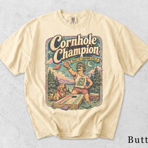 Könnte beinhalten: Ein butterfarbenes T-Shirt mit einer grafischen Illustration einer Person, die Cornhole spielt, mit einem Hund in der Nähe. Der Text lautet Cornhole Champion und Bags Fly, Legends Stay. Das Design zeigt eine Landschaft mit Bäumen und Bergen.