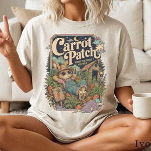 Può includere: Una t-shirt color avorio con la grafica di un coniglio con occhiali da sole e salopette, sdraiato in un giardino. Il testo recita Carrot Patch Where The Roots Are Wild. Il design include un piccolo capanno in legno, carote e alberi in stile retrò.