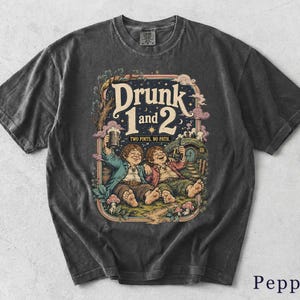 Peut inclure: Un t-shirt gris anthracite avec une illustration graphique de deux personnages assis dans l'herbe avec des chopes, entourés d'arbres et d'une petite maison. Le texte indique Drunk 1 and 2, Two Pints, No Path. Le t-shirt a un aspect délavé.