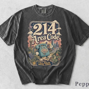 Könnte beinhalten: Ein anthrazitfarbenes T-Shirt mit einem Grafikdruck eines blauen Wählscheibentelefons mit Armen und Beinen. Der Text lautet 214 Area Code und Y'ALL DIALIN' GREAT TODAY. Das Design zeigt eine Skyline, Wolken, Sterne und Kakteen.