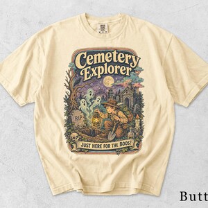 Könnte beinhalten: Ein butterfarbenes T-Shirt mit einer grafischen Illustration einer Person, die einen Friedhof mit Geistern, einer Laterne und einer Schaufel erkundet. Der Text lautet Cemetery Explorer und Just Here For The Boos, vor Grabsteinen.