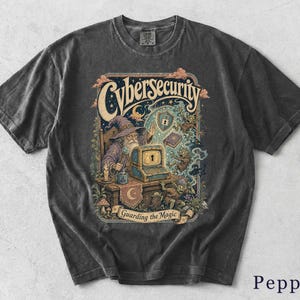 Può includere: Una t-shirt grigio antracite con un'illustrazione grafica di un mago seduto a una scrivania con un computer. Il testo "Cybersecurity" appare in alto e "Guarding the Magic" su uno stendardo in basso. Il design include uno scudo, un libro e piccole creature.