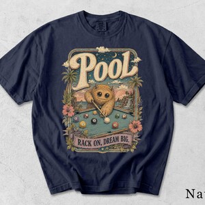 Könnte beinhalten: Ein marineblaues T-Shirt mit einer grafischen Illustration eines pelzigen Wesens, das an einem Billardtisch spielt. Das Design zeigt Palmen, Hibiskusblüten sowie den Text "POOL" oben und "RACK ON, DREAM BIG." unten im Rahmen.