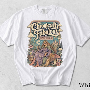 Może przedstawiać: Biały t-shirt z kolorowym nadrukiem graficznym z napisem "Chronically Fabulous" oraz "Pain is temporary, but the vibes are forever." Ilustracja przedstawia osobę w fioletowej szacie siedzącą na krześle z kotem.