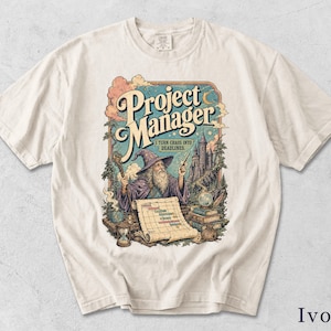 Könnte beinhalten: Ein elfenbeinfarbenes T-Shirt mit einer grafischen Illustration eines Zauberers in einer violetten Robe, der einen Stab und einen Zauberstab hält. Der Text lautet Project Manager und I TURN CHAOS INTO DEADLINES. Das Design zeigt ein Schloss, eine Schriftrolle, Bücher und eine Sanduhr.