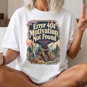 Könnte beinhalten: Ein weißes Kurzarm-T-Shirt mit einem farbenfrohen Grafikdruck mit dem Text "Error 404 Motivation Not Found" und "Please try napping and snacks". Die Abbildung zeigt eine Person, die mit gesenktem Kopf in der Nähe eines Schwertes sitzt.