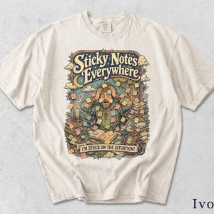 May include: An ivory short-sleeve t-shirt featuring a colorful graphic of a person surrounded by stacks of books and numerous sticky notes. The text reads Sticky Notes Everywhere and I'M STUCK ON THE SITUATION! The design includes a candle, a quill, and a starry night sky background.