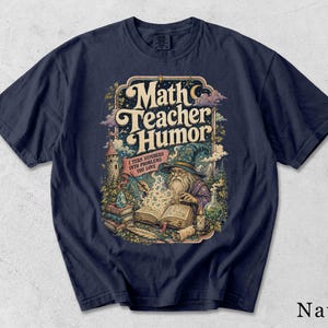 Può includere: Una t-shirt blu navy con un'illustrazione grafica di un mago che legge un libro con numeri fluttuanti. Il testo recita "Math Teacher Humor" e "I turn numbers into problems you love." Il design include un castello, alberi e un cristallo.