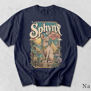 Może przedstawiać: Granatowy t-shirt z nadrukiem graficznym przedstawiającym kota rasy sfinks siedzącego przed egipskimi piramidami. Napis głosi "Sphynx" oraz "No fur. All attitude.". Ilustracja zawiera palmy, półksiężyc oraz ozdobny wazon.