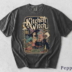 Könnte beinhalten: Ein anthrazitfarbenes Kurzarm-T-Shirt mit einem Grafikdruck einer Person mit Hexenhut, die in einem Kessel rührt. Der Text lautet Kitchen Witch und Stirring Up Trouble & Soup. Die Illustration zeigt zudem eine schwarze Katze und einen Mond.