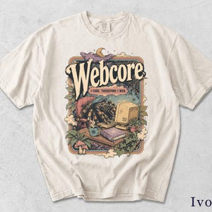 May include: An ivory t-shirt featuring a graphic print of a spider sitting at a desk with a computer, a mug, and a book. The text reads Webcore and I CODE, THEREFORE I WEB. The design includes a crescent moon, spiderwebs, and foliage elements.