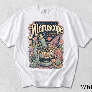 Może przedstawiać: Biały t-shirt z kolorowym nadrukiem graficznym przedstawiającym czarodzieja patrzącego przez mikroskop. Tekst brzmi Microscope Big Adventures In Tiny Places. Wzór zawiera planetę, szklaną kolbę, książkę oraz małe stworzenia w kosmicznej scenerii.