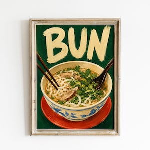 Könnte beinhalten: Ein gerahmter Kunstdruck mit einer Schüssel Nudelsuppe, garniert mit grünen Kräutern und Fleischstücken. Das Wort BUN steht in großen, hellgelben Pinselstrich-Buchstaben darüber. Die Schüssel hat ein blaues Blumenmuster und steht auf einem roten Teller.
