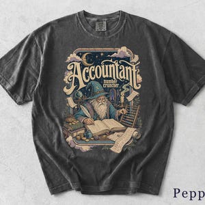 Könnte beinhalten: Ein anthrazitfarbenes T-Shirt mit einer Grafik eines Zauberers, der in ein Buch schreibt, neben einem Abakus, Münzen und Schriftrollen. Der Text lautet Accountant number cruncher. Das Design zeigt einen Halbmond, Sterne und Wolken im Rahmen.
