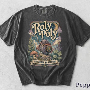 Może przedstawiać: Ciemnoszary t-shirt z grafiką przedstawiającą prosionka przebranego za rycerza z mieczem i tarczą. Napis głosi Roly Poly oraz Tiny Armor, Big Attitude. Wzór zawiera grzyby, rośliny, chmury oraz sierp księżyca w tle.