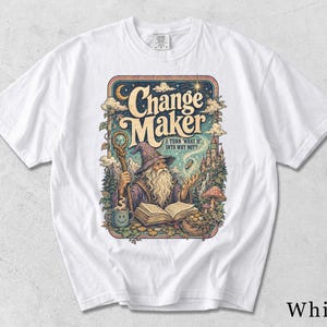 May include: A white short-sleeve t-shirt featuring a colorful graphic print of a wizard with a staff, an open book, and a castle. The text reads Change Maker and I TURN WHAT IF INTO WHY NOT? The design includes clouds, stars, and a mushroom.