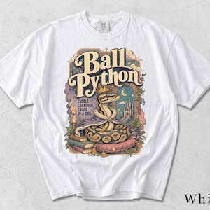 Könnte beinhalten: Ein weißes T-Shirt mit einer grafischen Illustration eines Königspythons, der eine goldene Krone und einen violetten Umhang trägt und auf einem rosa Kissen liegt. Der Text lautet Ball Python, Cuddle Champion, Chaos In A Coil, mit Schloss und Büchern.