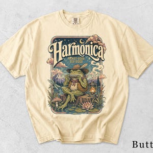 May include: A butter-colored t-shirt featuring a graphic of a frog wearing a hat and vest, playing a harmonica while sitting on a log. The text reads "Harmonica" and "Pocket-sized, ego-sized." The scene includes a lantern, mountains, and a dragonfly.