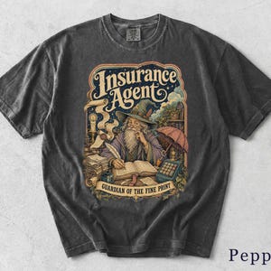 Könnte beinhalten: Ein anthrazitfarbenes T-Shirt mit einer grafischen Illustration eines Zauberers, der in ein Buch schreibt. Der Text lautet Insurance Agent und Guardian of the Fine Print. Das Design umfasst eine Kerze, eine Schriftrolle, einen Taschenrechner und einen Regenschirm, die alle in einem detaillierten, farbenfrohen Stil auf der Vorderseite des Kleidungsstücks dargestellt sind.
