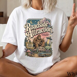 May include: A white t-shirt featuring a graphic print with the text "Roadside Attraction" and "Because normal is overrated." The illustration depicts a brown Bigfoot, a green dinosaur, a flying saucer, a station wagon, and a "World's Okayest Giant" sign.