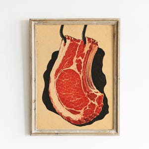Könnte beinhalten: Ein gerahmter Kunstdruck mit einer grafischen Illustration eines rohen Koteletts mit Knochen, das an zwei schwarzen Haken hängt. Das Fleisch ist in leuchtenden Rot- und Rosatönen dargestellt, vor einer schwarzen Silhouette auf beigem Grund.