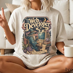 Könnte beinhalten: Ein elfenbeinfarbenes T-Shirt mit einem Grafikdruck eines Zauberers, der an einem Schreibtisch mit Computer, Büchern und einer Tasse sitzt. Der Text lautet Web Developer und Debugging the Arcane. Das Bild zeigt Sterne und einen Mond.