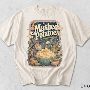 May include: An ivory t-shirt featuring a graphic print of a wizard holding a staff, a bowl of mashed potatoes with butter, a gravy boat, and a castle. The text reads "Mashed Potatoes" and "The original cloud food" in a stylized, vintage-inspired font.