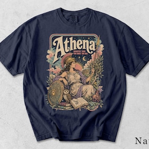 May include: A navy blue t-shirt featuring a graphic print of Athena with a helmet, spear, shield, owl, and books. The text reads Athena Smarter Than You Since 700 BCE. The design includes clouds, a sun, a moon, and a temple in a colorful palette.