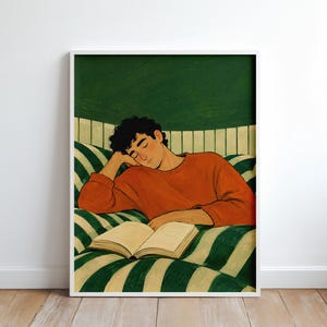 Könnte beinhalten: Ein gerahmter Kunstdruck mit einer Illustration einer Person mit dunklem, lockigem Haar, die einen orangefarbenen Pullover trägt, den Kopf auf eine Hand stützt und auf einer grün-weiß gestreiften Fläche liegt, vor der ein offenes Buch liegt.