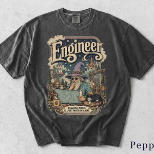Könnte beinhalten: Ein anthrazitfarbenes Kurzarm-T-Shirt mit einem Grafikdruck eines bärtigen Zauberers mit Hut, der einen Bauplan betrachtet. Der Text lautet "Engineer" und "BECAUSE MAGIC IS JUST MATH IN A HAT." Das Design zeigt eine Rakete, eine Burg und eine Katze.