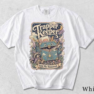 May include: A white t-shirt featuring a colorful graphic print of a blue, star-patterned binder with eyes, a raccoon wearing a wizard hat, and the text "Trapper Keeper" and "Hold My Homework" surrounded by clouds, stars, and school supplies.